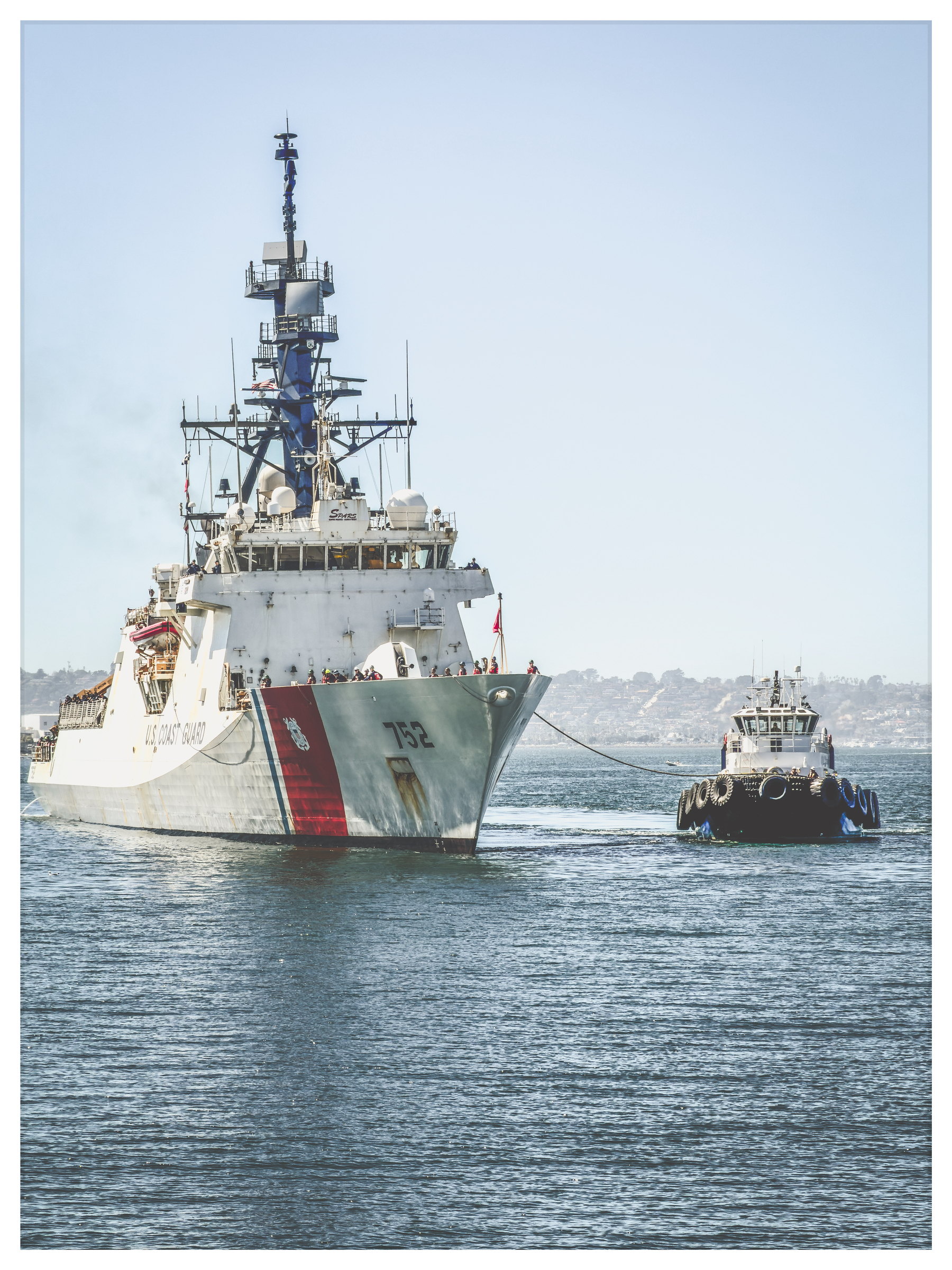 Auto-generated description: A large Coast Guard ship is being guided by a smaller tugboat on a calm body of water.