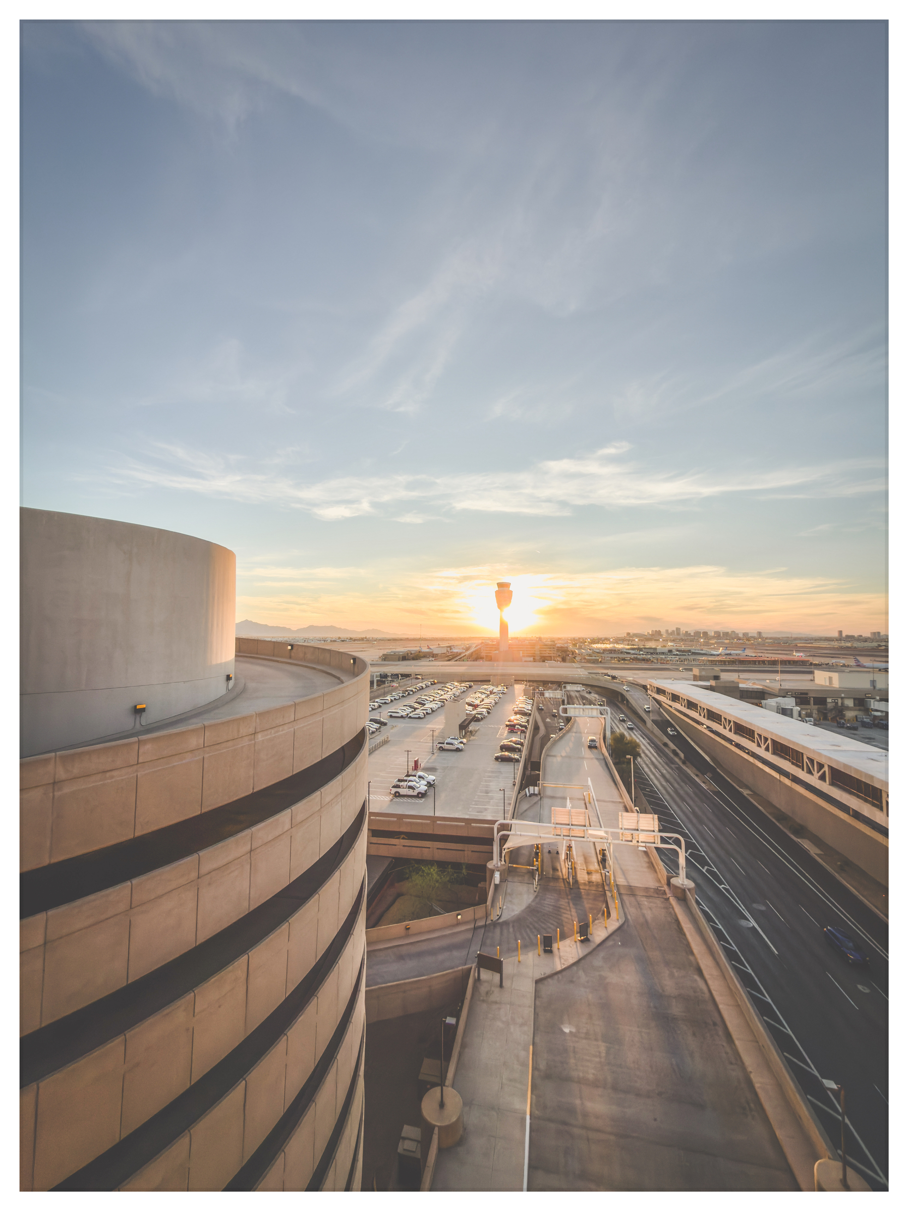 Auto-generated description: A sunset view over an airport, featuring an air traffic control tower, parking structures, and multiple runways and roads.