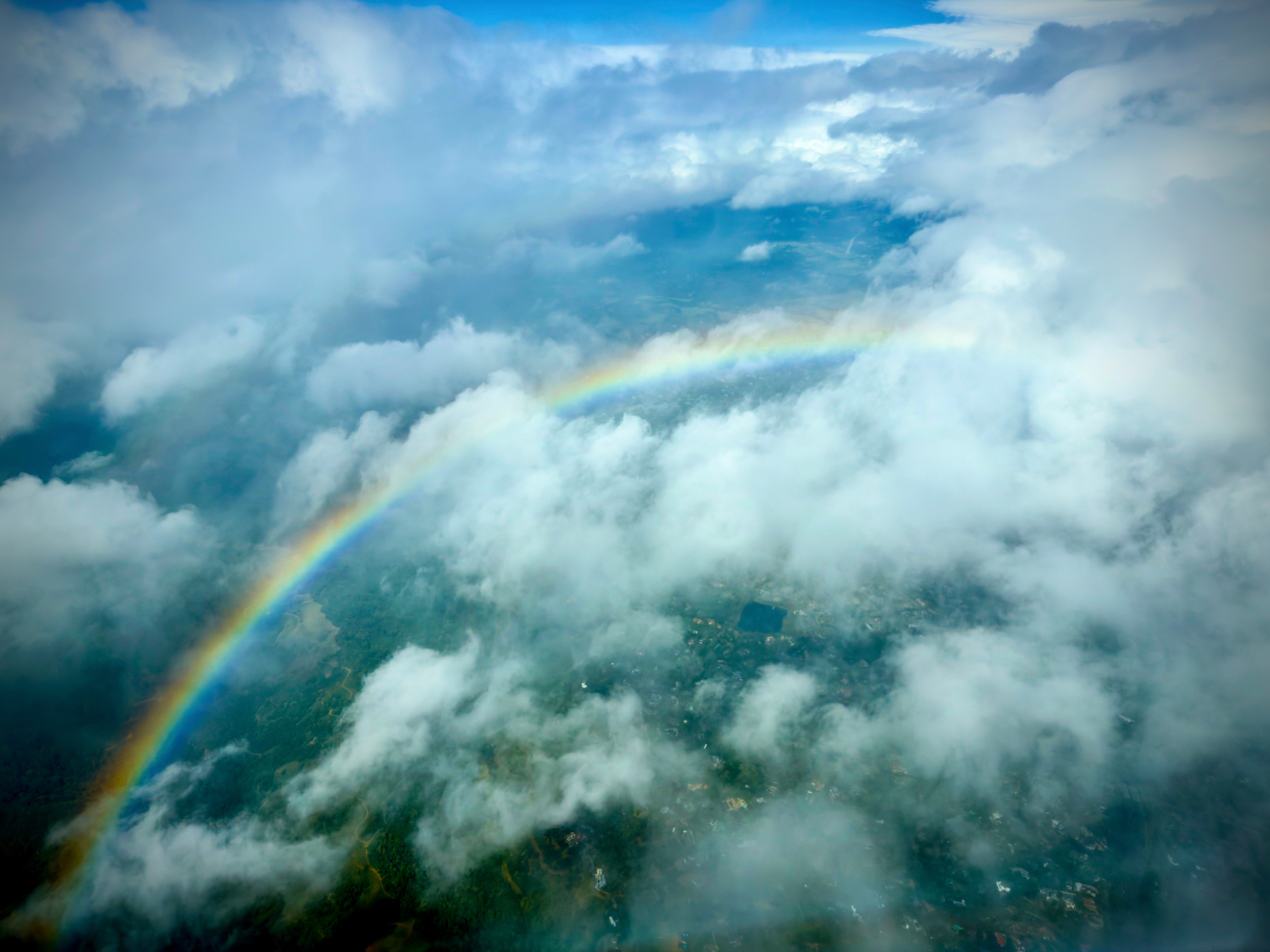 Auto-generated description: An aerial view features a vibrant rainbow arching over a landscape partially obscured by fluffy clouds.